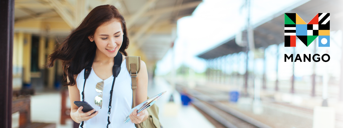 A person with long brown hair looks at a phone while standing on a train platform.