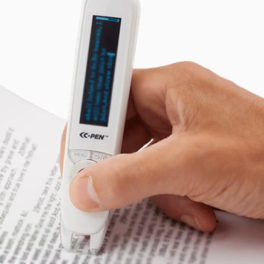 A person's hand holds a C-Pen Reader over text on an open book. 