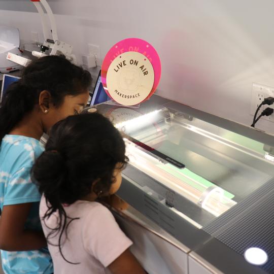 Two girls watch a laser cutter at work. 