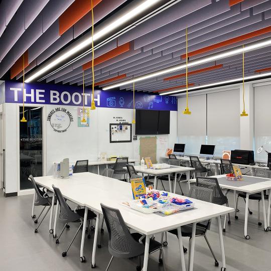 Interior of the Makerspace at McLean Branch. Long white tables and chairs with a blue sign in the background that says The Booth.