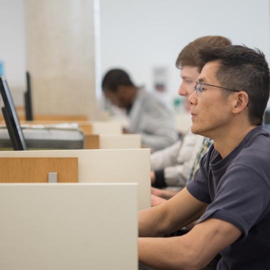 A man in a blue shirt, sitting at a computer, viewed from the side. 