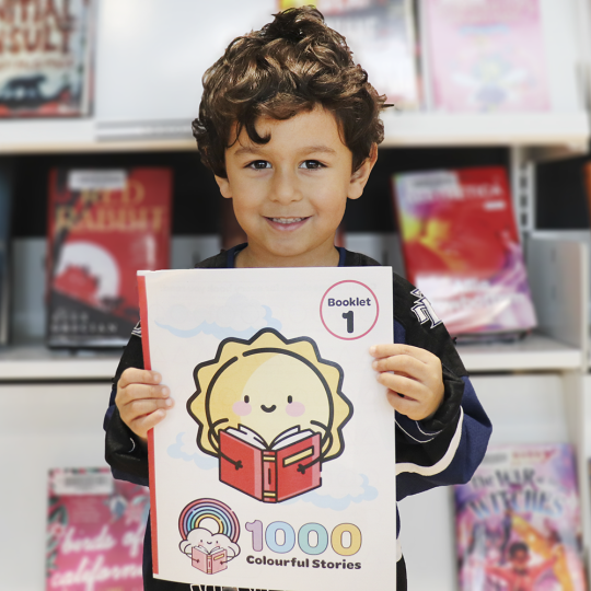 A child holds up a booklet with a smiling sun holding a red book on the cover. The child is standing in front of a book shelf at Audley Branch.