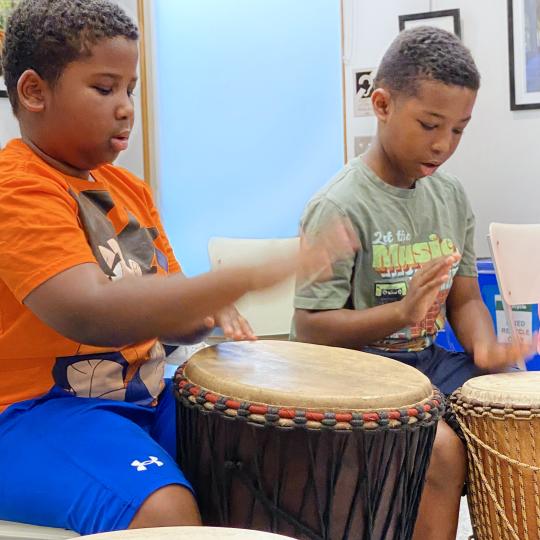 Two young boys sit in the Library playing drums as part of a summer program.