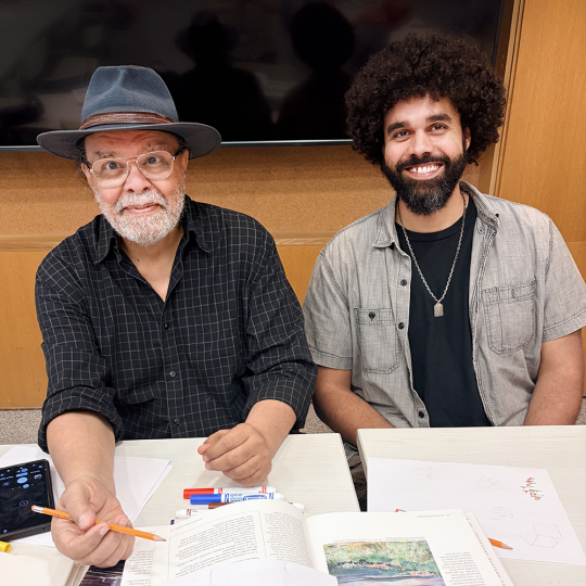 Two people sit together at a table in Main Branch with drawing supplies and books in front of them. They are smiling.